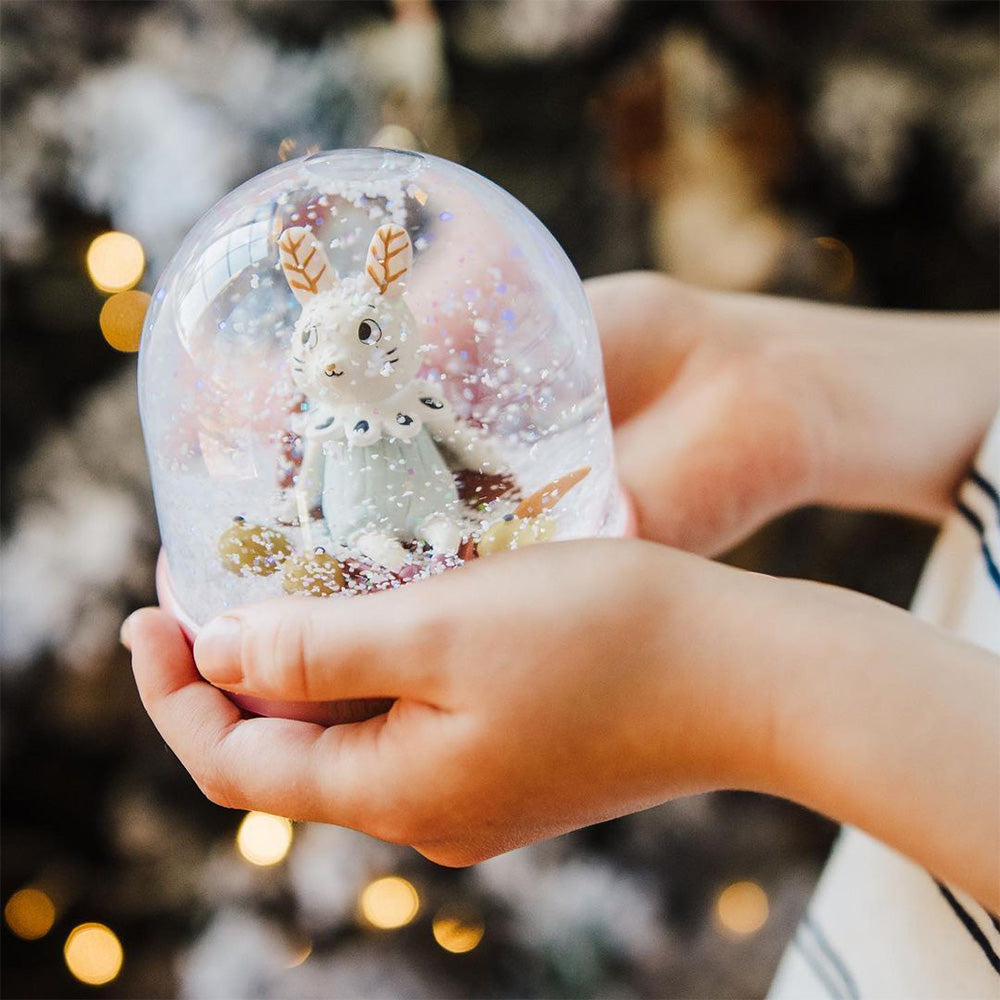 Hands holding Moulin Roty snow globe showing white rabbit in blue dress inside glass dome