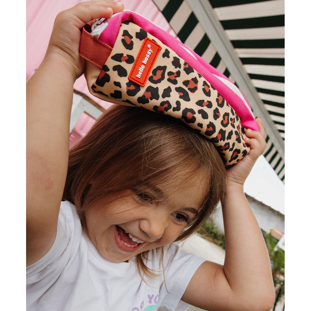 Child playfully holding Hello Hossy leopard print pencil case like a hat outdoors