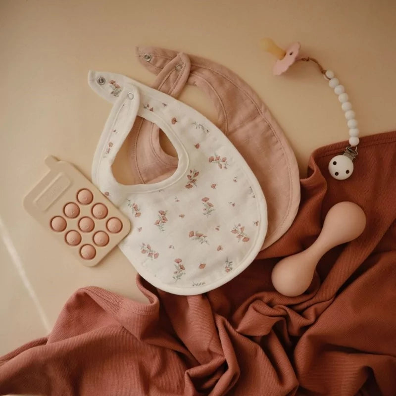 Stylized flat lay of MUSHIES floral and blush muslin bibs with wooden teether accessories