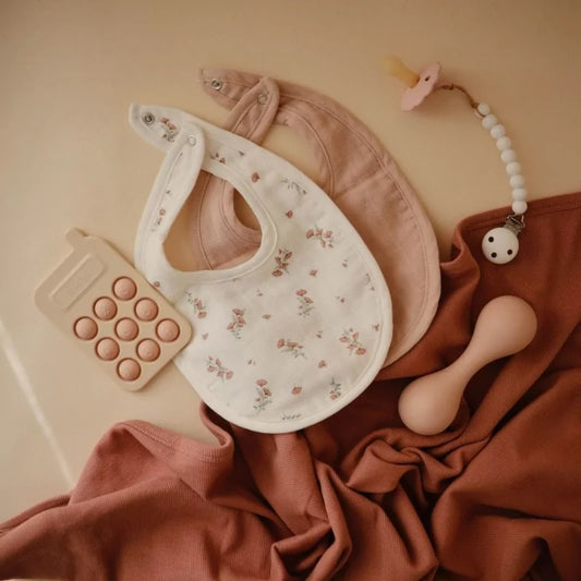 Stylized flat lay of MUSHIES floral and blush muslin bibs with wooden teether accessories