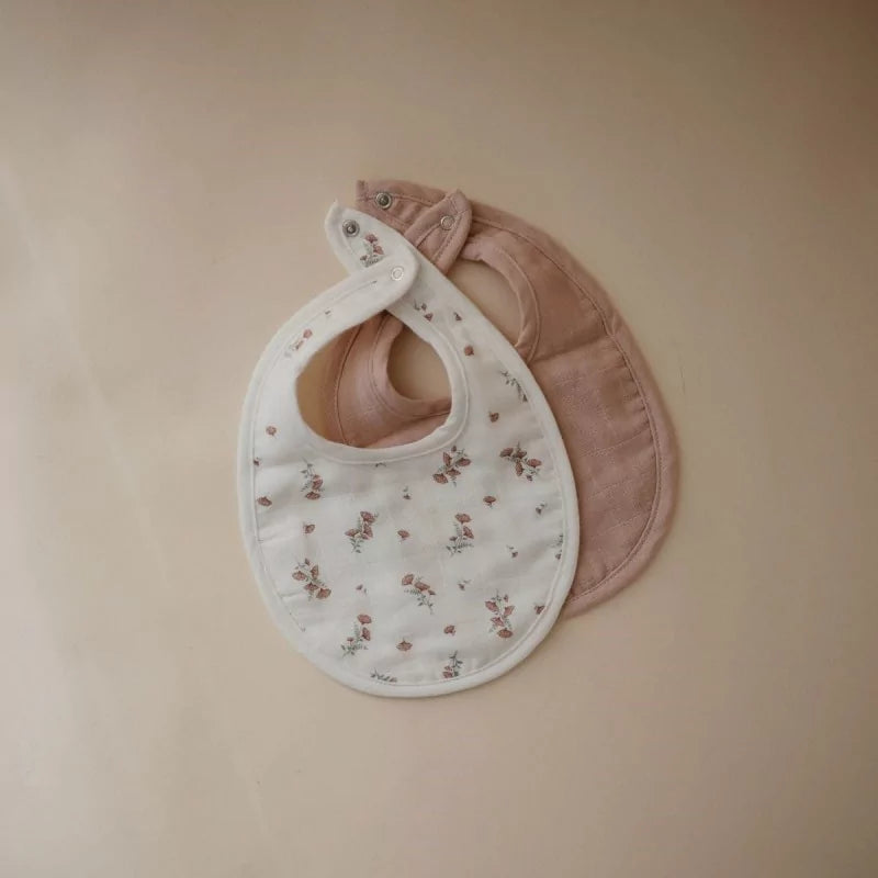 Flat lay of white floral and solid dusty blush muslin baby bibs showing silver snaps