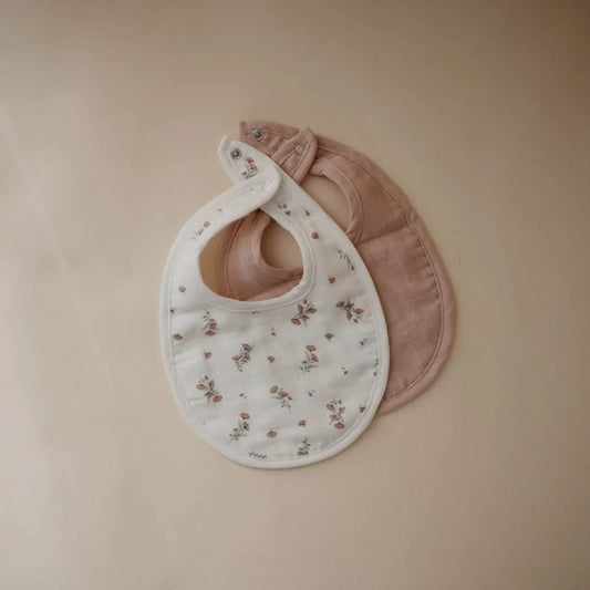 Flat lay of white floral and solid dusty blush muslin baby bibs showing silver snaps