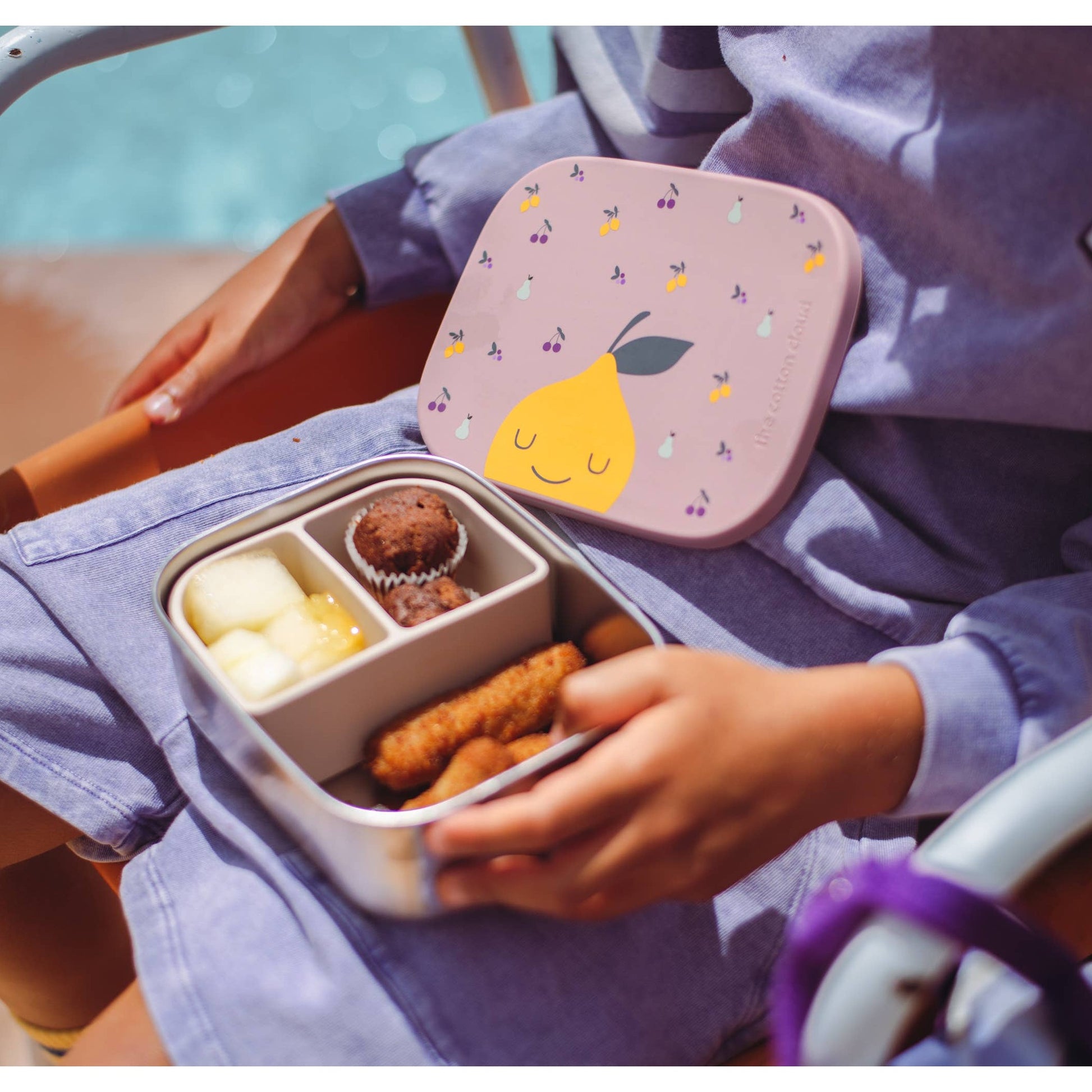 Child holding open pear-themed stainless steel lunchbox outdoors