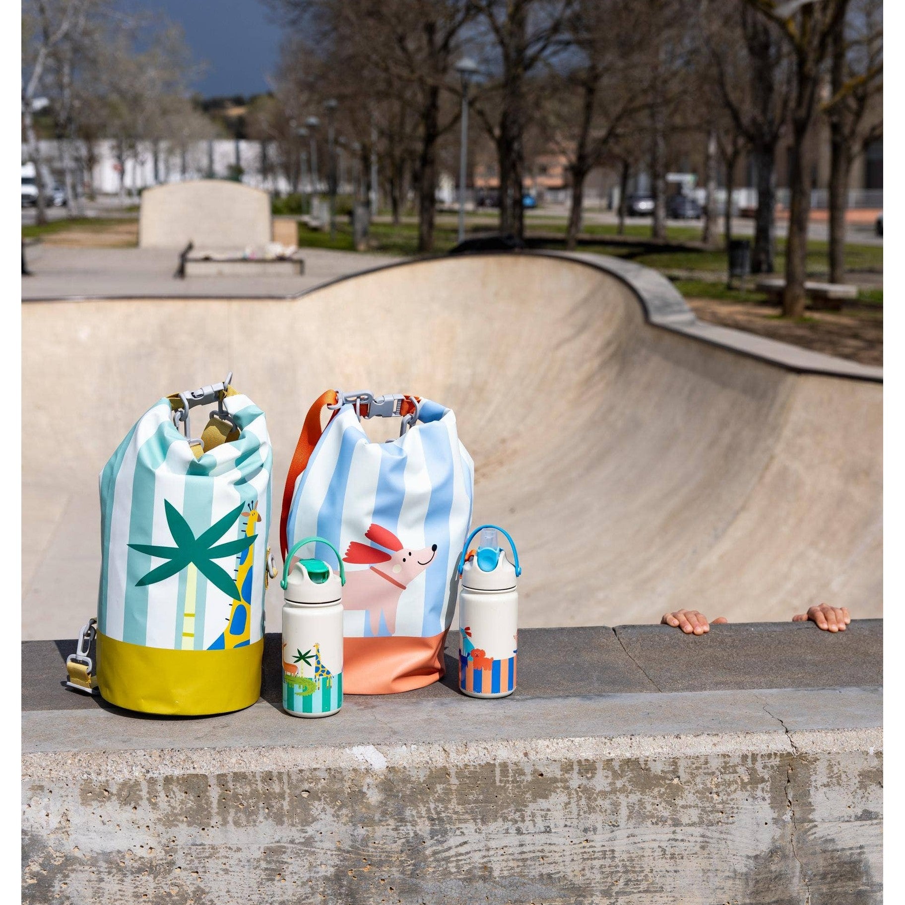 Kids insulated bottles with giraffe and dog graphics beside dry bags at skatepark