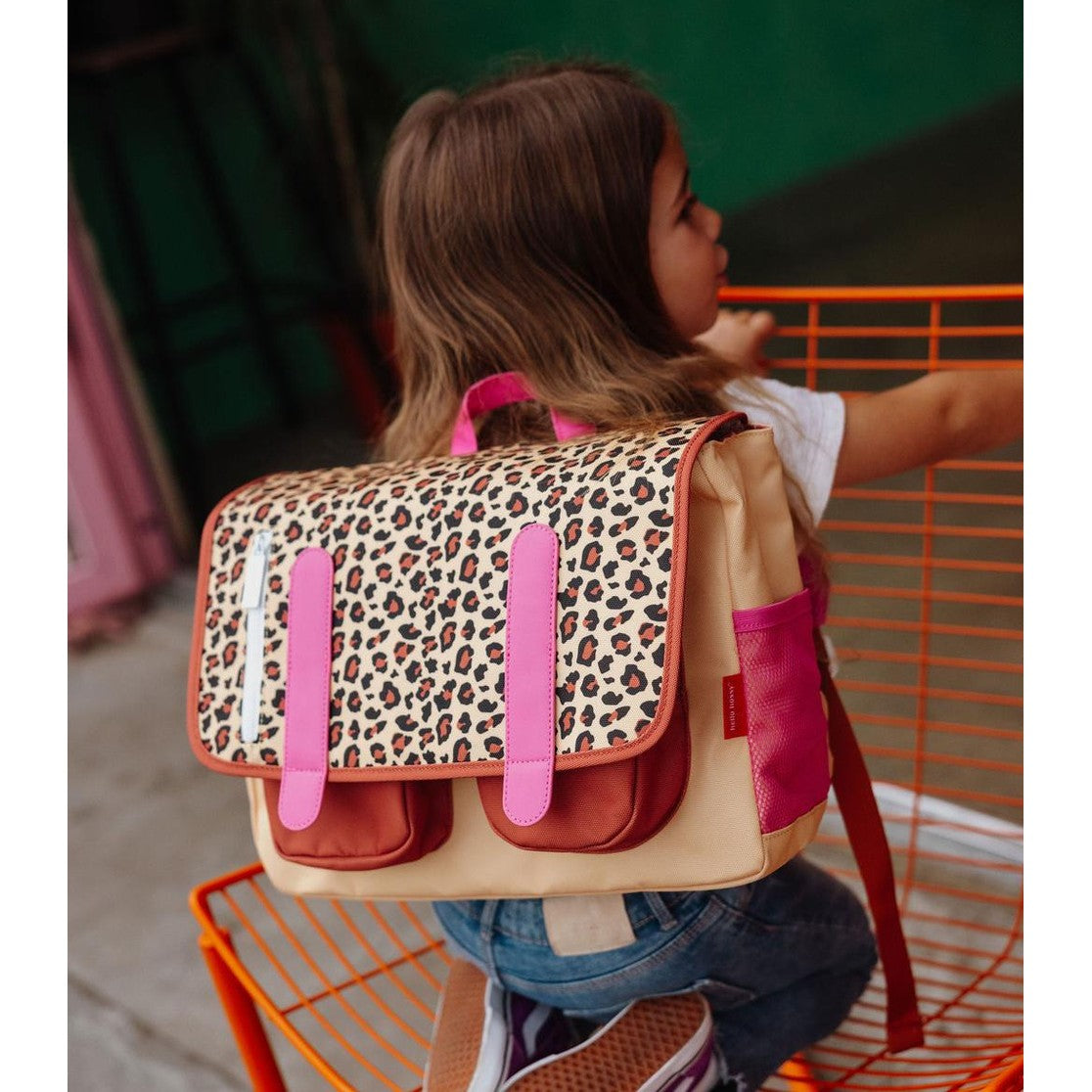 Child sitting on orange chair wearing beige leopard print school bag