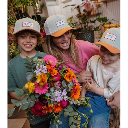 Mom and two boys in green and orange Hello Hossy kids trucker hats near flowers