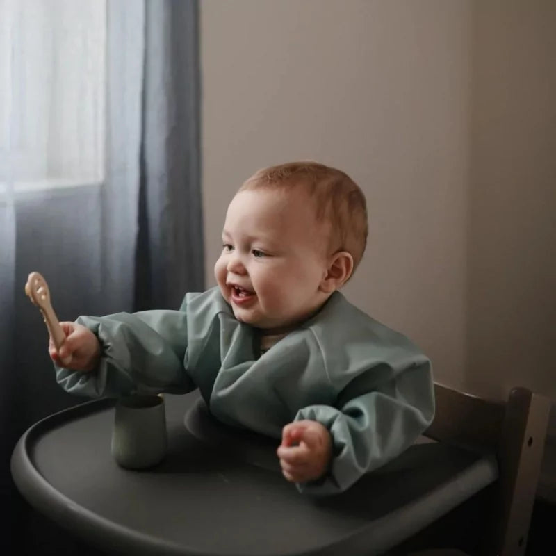Baby holding beige MUSHIES silicone feeding spoon while sitting in high chair with matching sage green bib