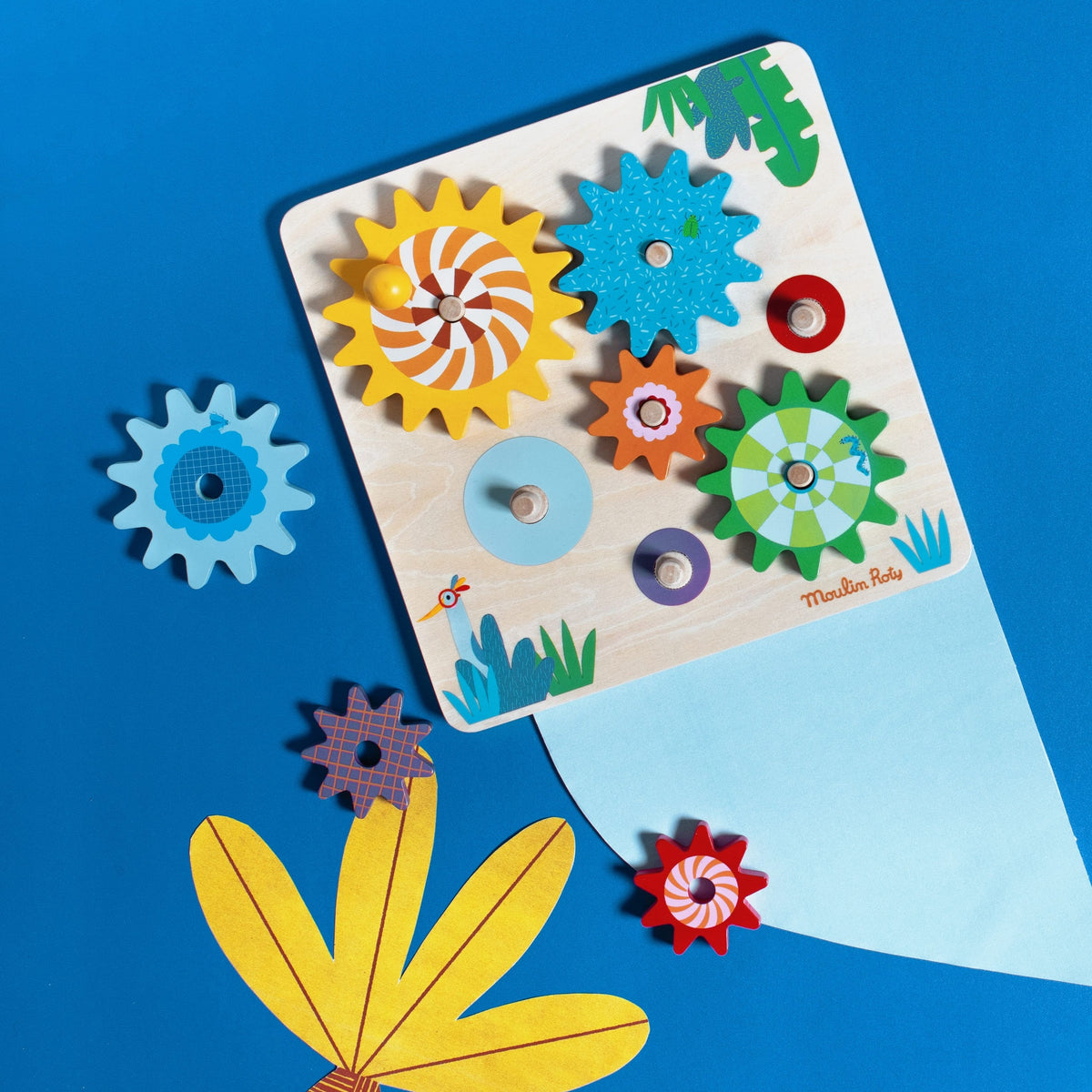 Angled view of Moulin Roty wooden gear board with attached and loose colorful gears on blue background