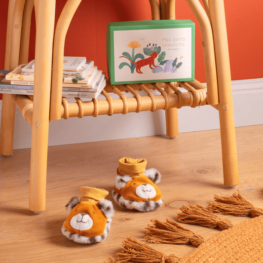 Orange and grey animal face baby slippers on wooden floor near rattan chair