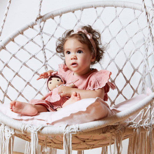 Toddler holding Kaloo Amandine plush doll while sitting in macrame chair