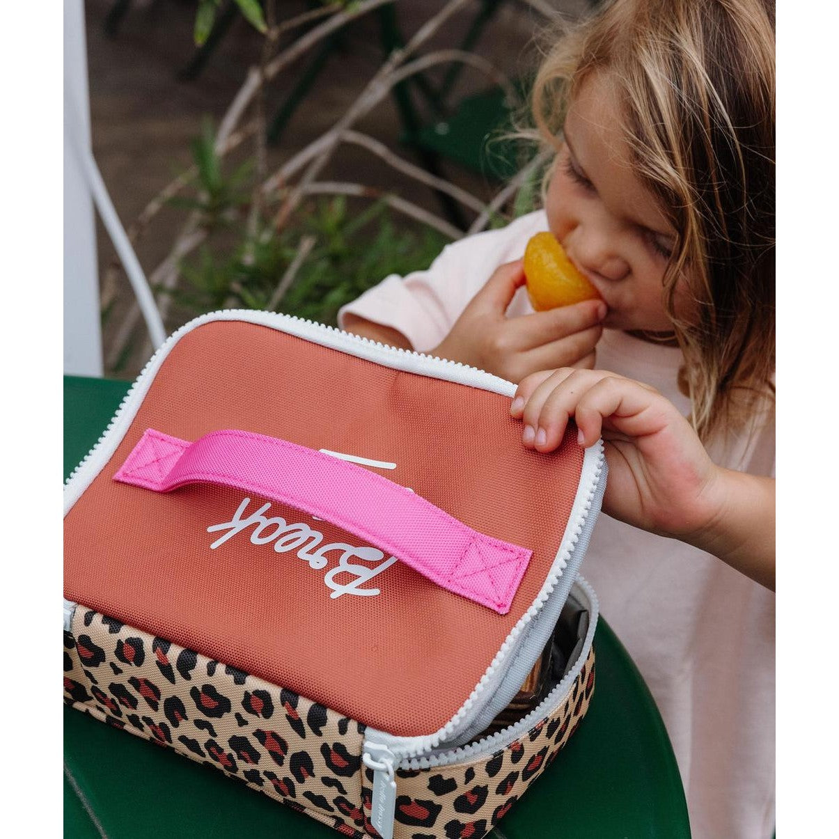 Child biting fruit next to open leopard print lunch bag on green surface
