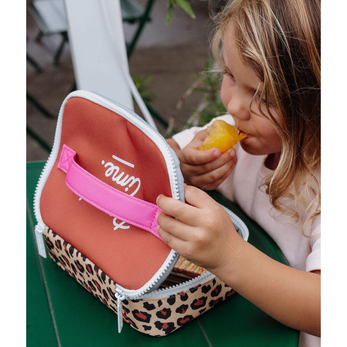 Child holding open leopard print lunch bag while eating frozen treat outdoors