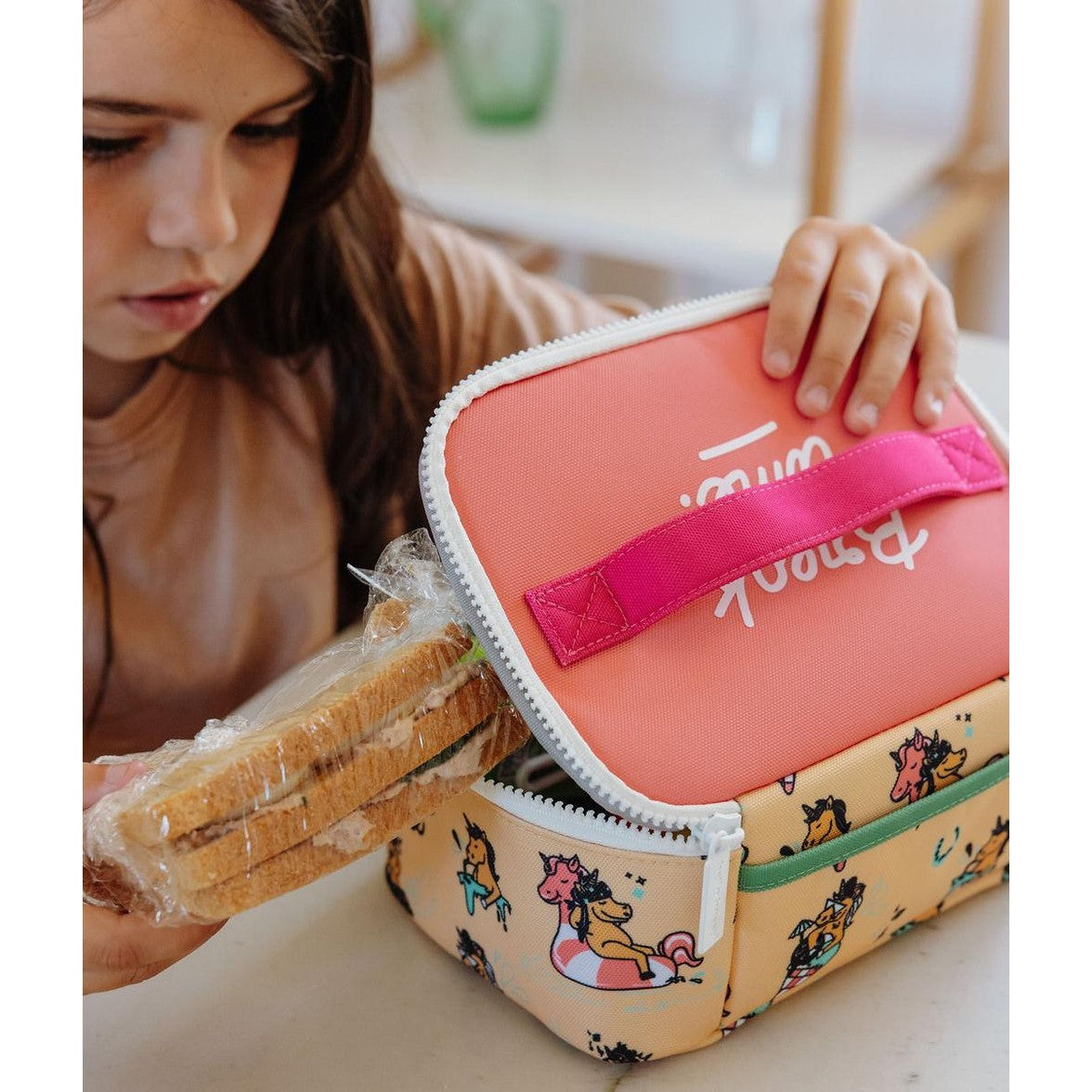 Child holding beige unicorn print insulated lunch bag with coral lid