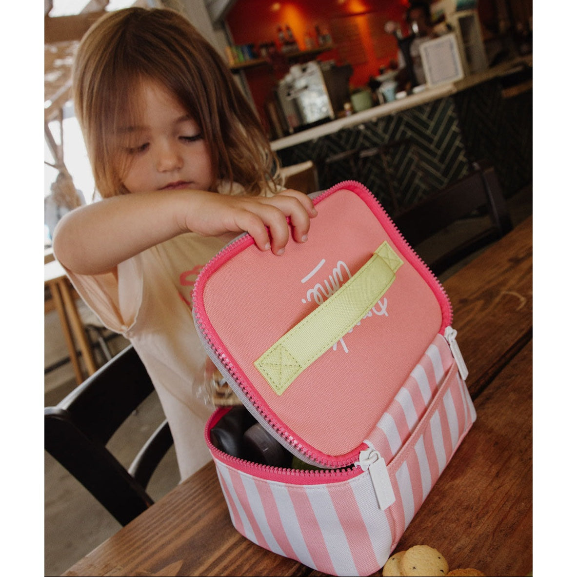Child opening pink striped insulated lunch bag in café setting