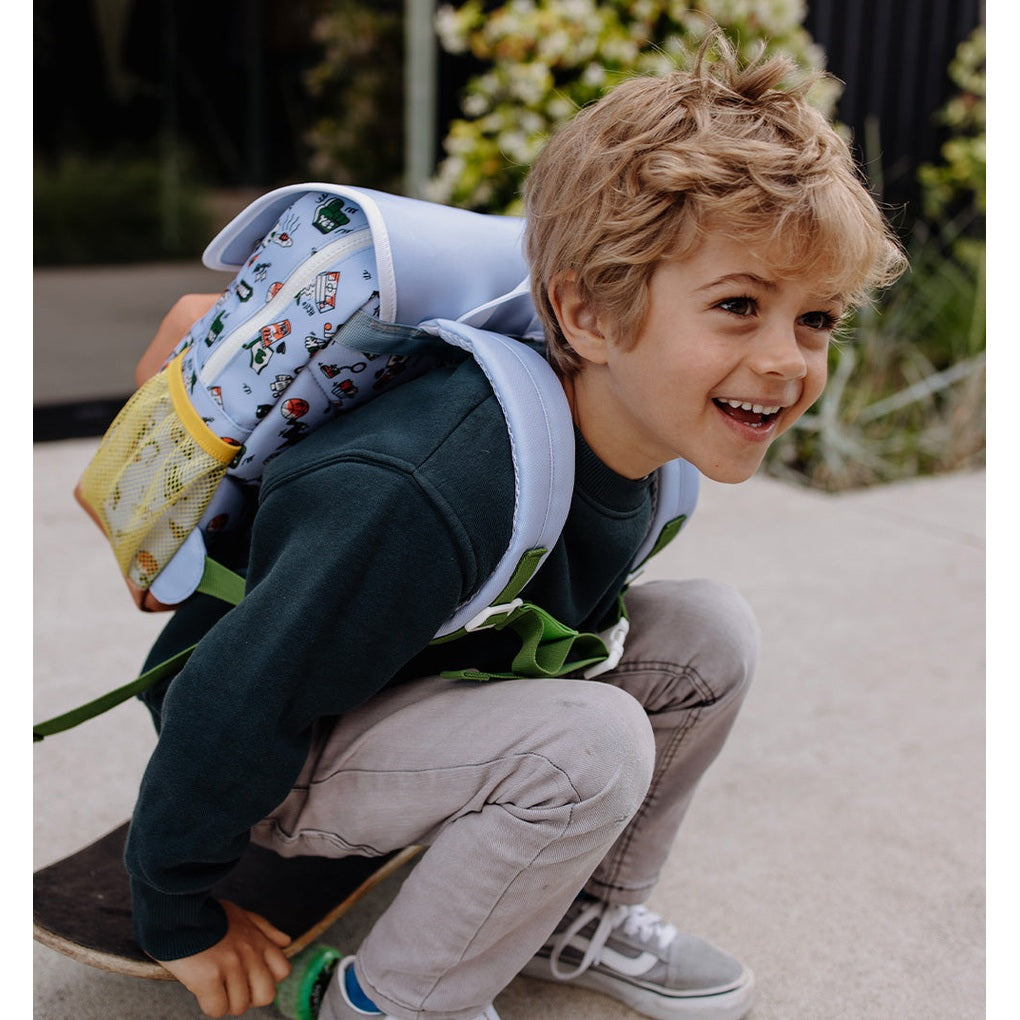 Smiling child in green sweater wearing light blue Dunk backpack on skateboard