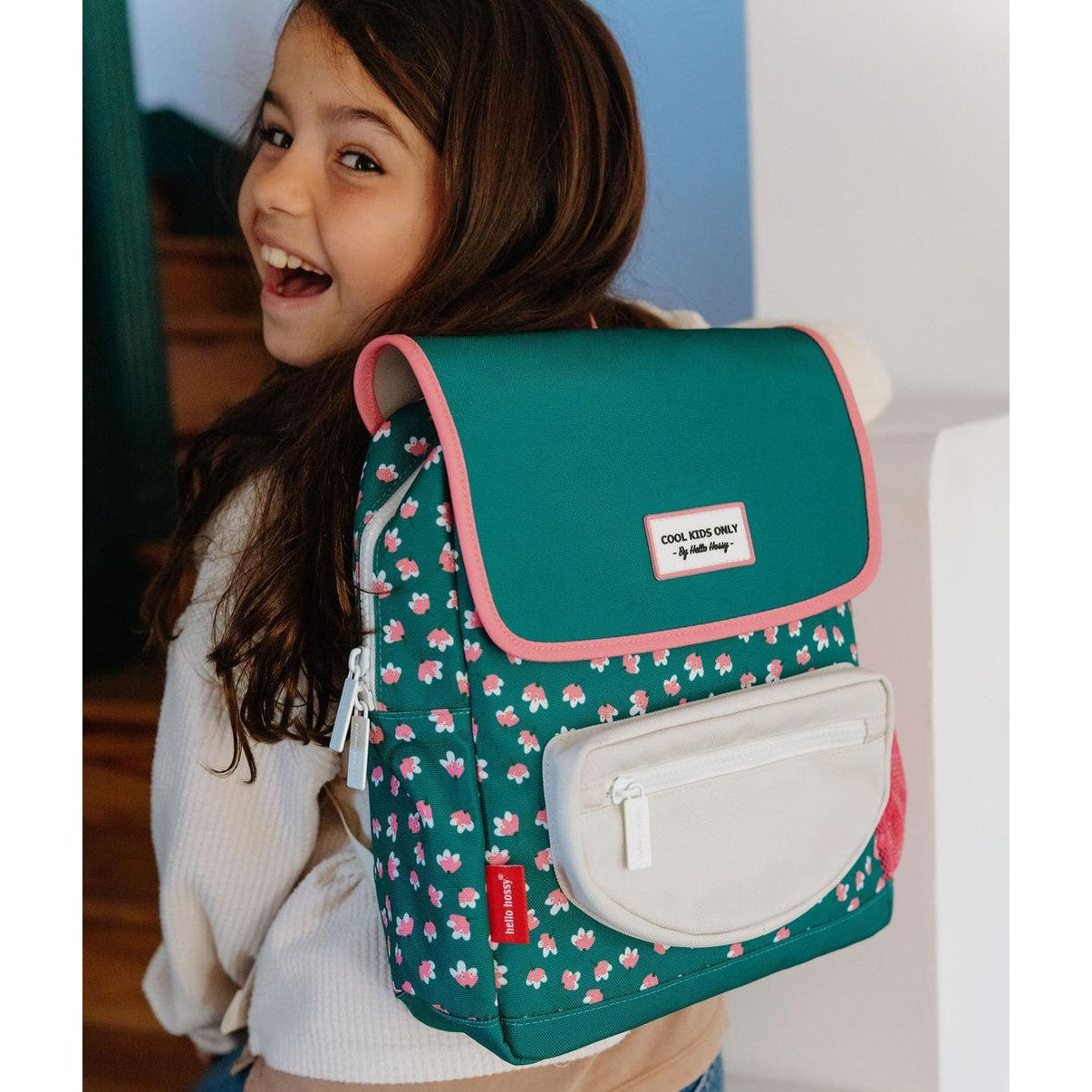 Girl smiling while wearing green floral square backpack with Hello Hossy tag