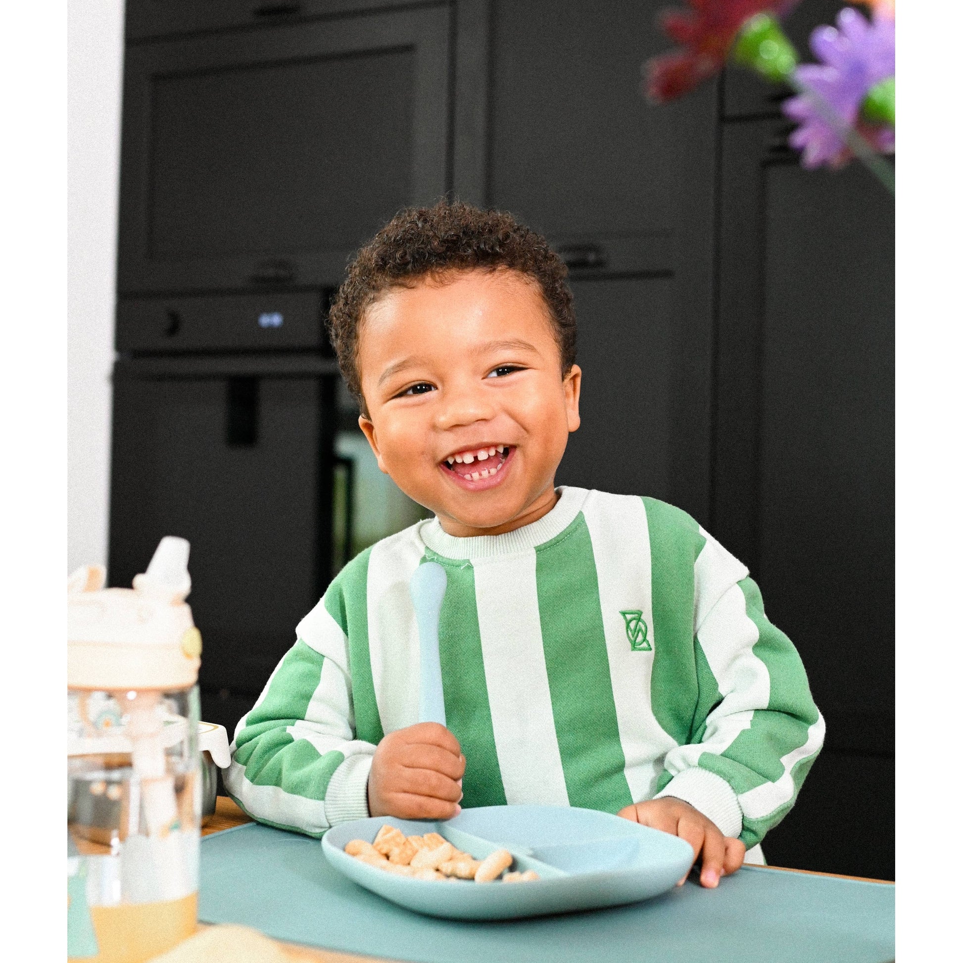 Smiling toddler eating snacks from blue divided plate with silicone spoon