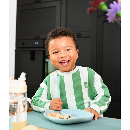 Smiling toddler eating snacks from blue divided plate with silicone spoon