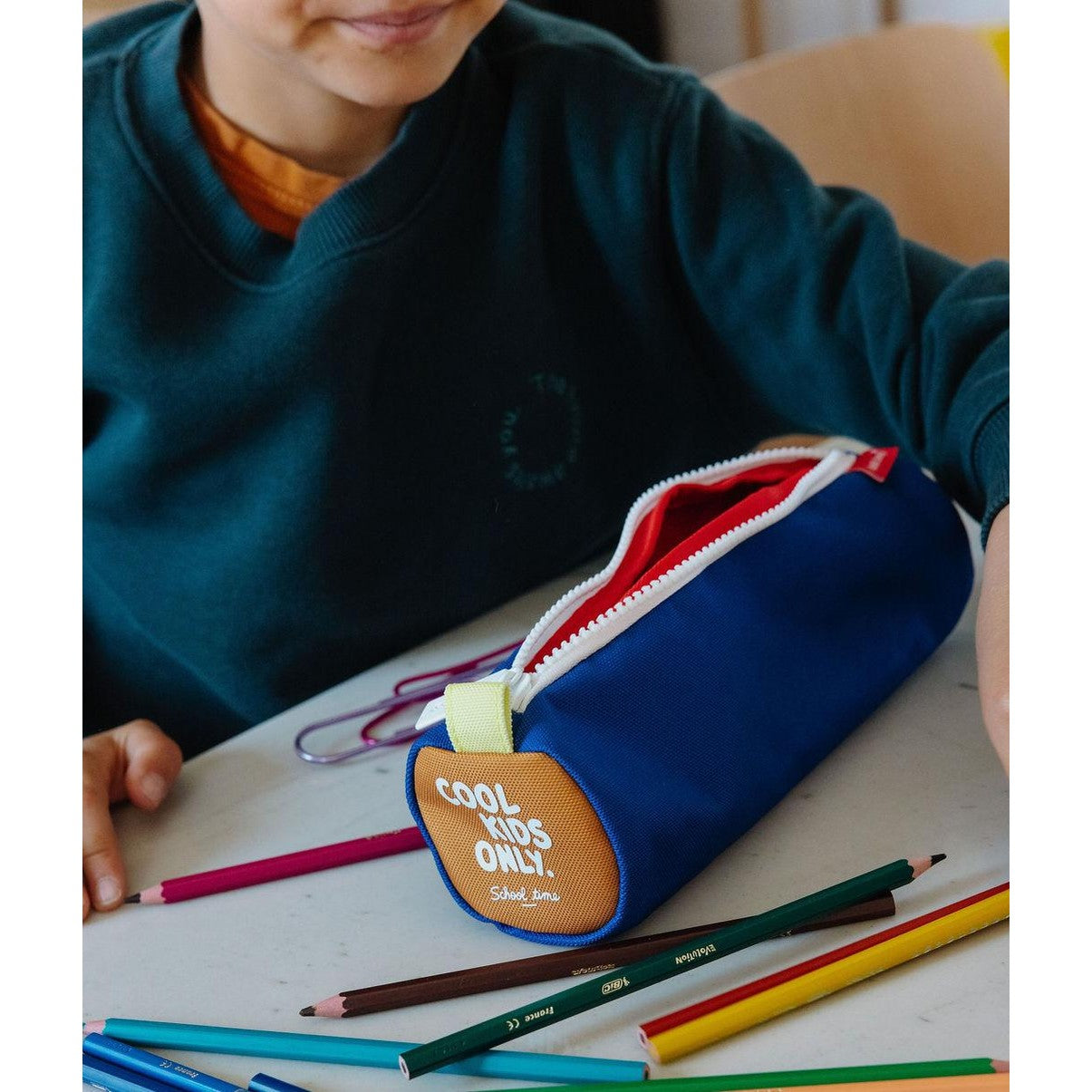 Blue kids stationery bag with red lining surrounded by colored pencils on desk